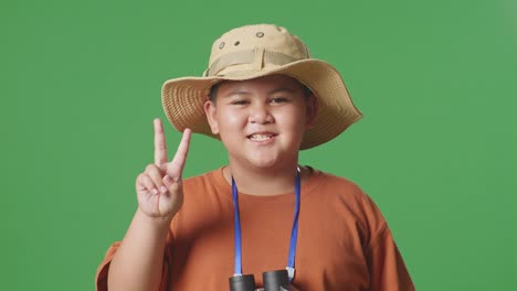 niño turista asiático con un sombrero y binoculares usando la lupa luego mostrando el gesto de paz en el fondo de la pantalla verde. niño investigador examina algo, aventura de turismo de viaje, de cerca