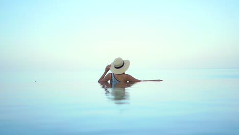 while resting on the edge of an infinity pool, a woman with her back to the camera gives the illusion of the water from the pool and ocean are one