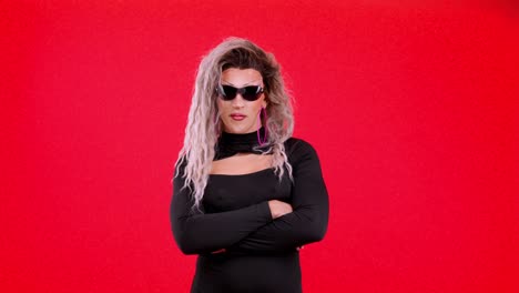 a confident drag queen in a black dress and sunglasses posing against a red background.