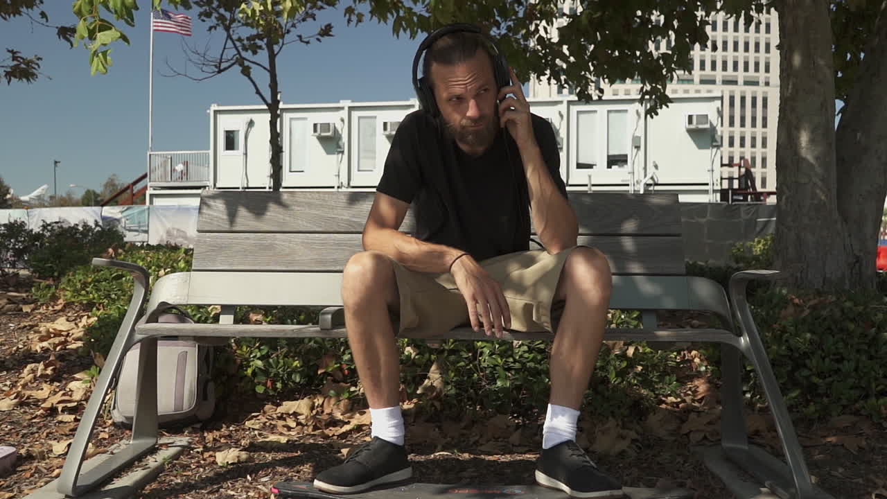 Stylish young adult male with skateboard sitting on park bench listening to music in headphones (Cinematic Slow Motion)