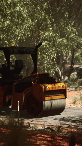 a yellow road roller is shown working on a dirt road in a forest. the road roller is moving forward and there is a small section of black tarmac in front of it. there are trees and bushes on either side of the road, and the road is surrounded by dry, red earth.  the image is taken from a slightly lo