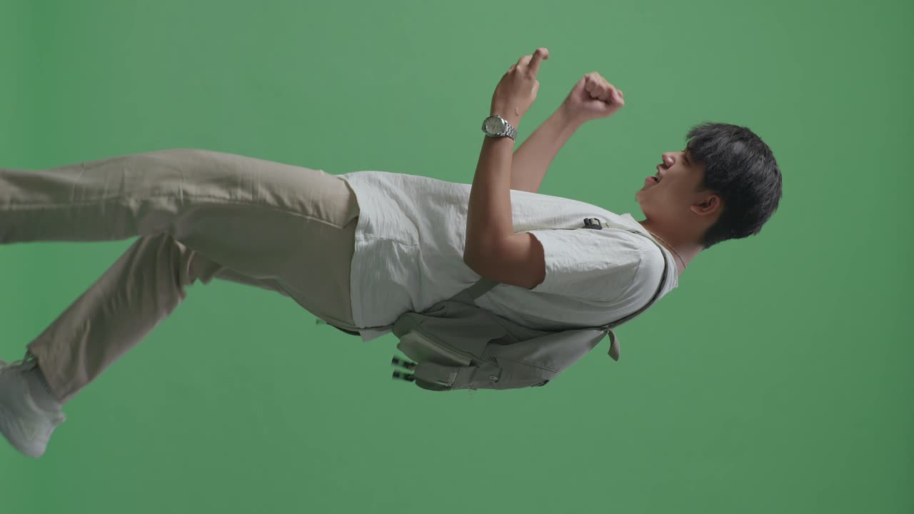 vista lateral de un estudiante asiático feliz jugando a un juego móvil y ganando mientras camina en el estudio de pantalla verde