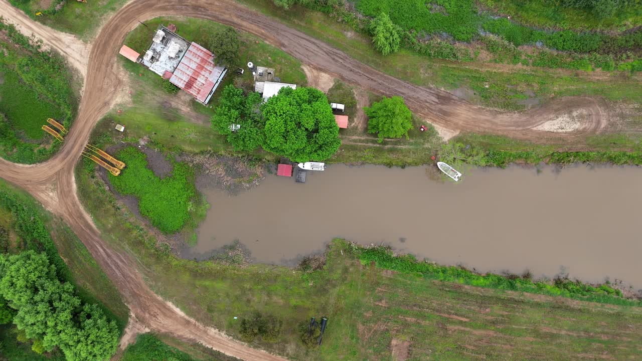 A high-angle aerial drone shot looking straight down at simple rural homes with tin roofs, boats, and dirt roads along a muddy canal in the Delta region of Argentina