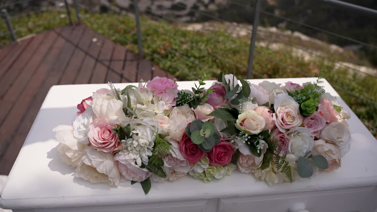Beautiful flowers are artfully arranged on a white table at an outdoor location. This colorful display merges roses, peonies, and greens, enhancing the scenic background