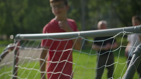 a soccer ball is shot across a goal post as the goalkeeper in red attempts to save it, while two people watch in the blurred background