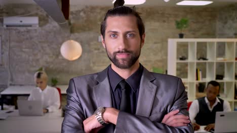 handsome businessman with beard is standing with arms crossed and watching at camera in office, colleagues are working with laptops, work concept