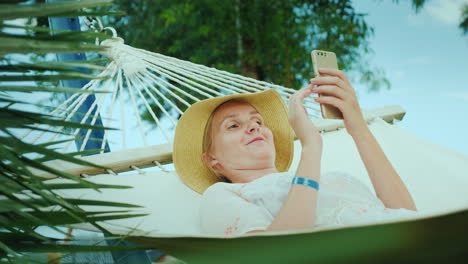Retrato-De-Una-Mujer-De-Vacaciones-Yace-En-Una-Hamaca-Utiliza-Un-Teléfono-Inteligente-4k-Video