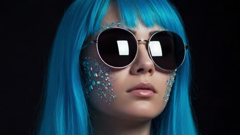 captivating expression: a fashion-forward individual with vibrant blue hair and striking makeup, posing elegantly in sunglasses against a dark background