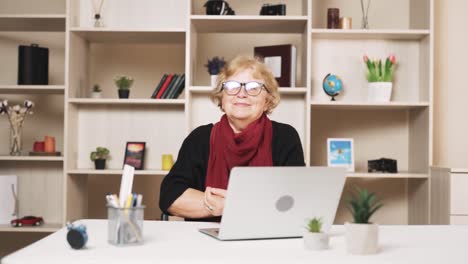 the older woman in glasses puts on glasses with a smile, looking into the camera while sitting at a table with a laptop