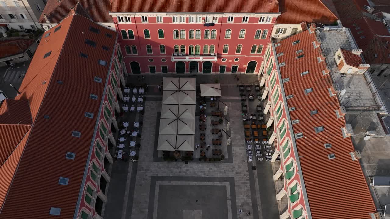 A lively plaza with tables under umbrellas surrounded by red rooftops, aerial view