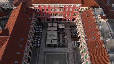 a lively plaza with tables under umbrellas surrounded by red rooftops, aerial view