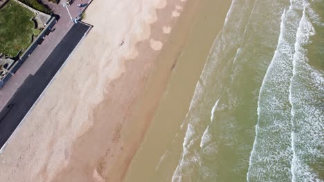 aerial footage that starts over waves on a sandy beach then rotates around to be looking down over the beach and garden promenade on weymouth beach, dorset.