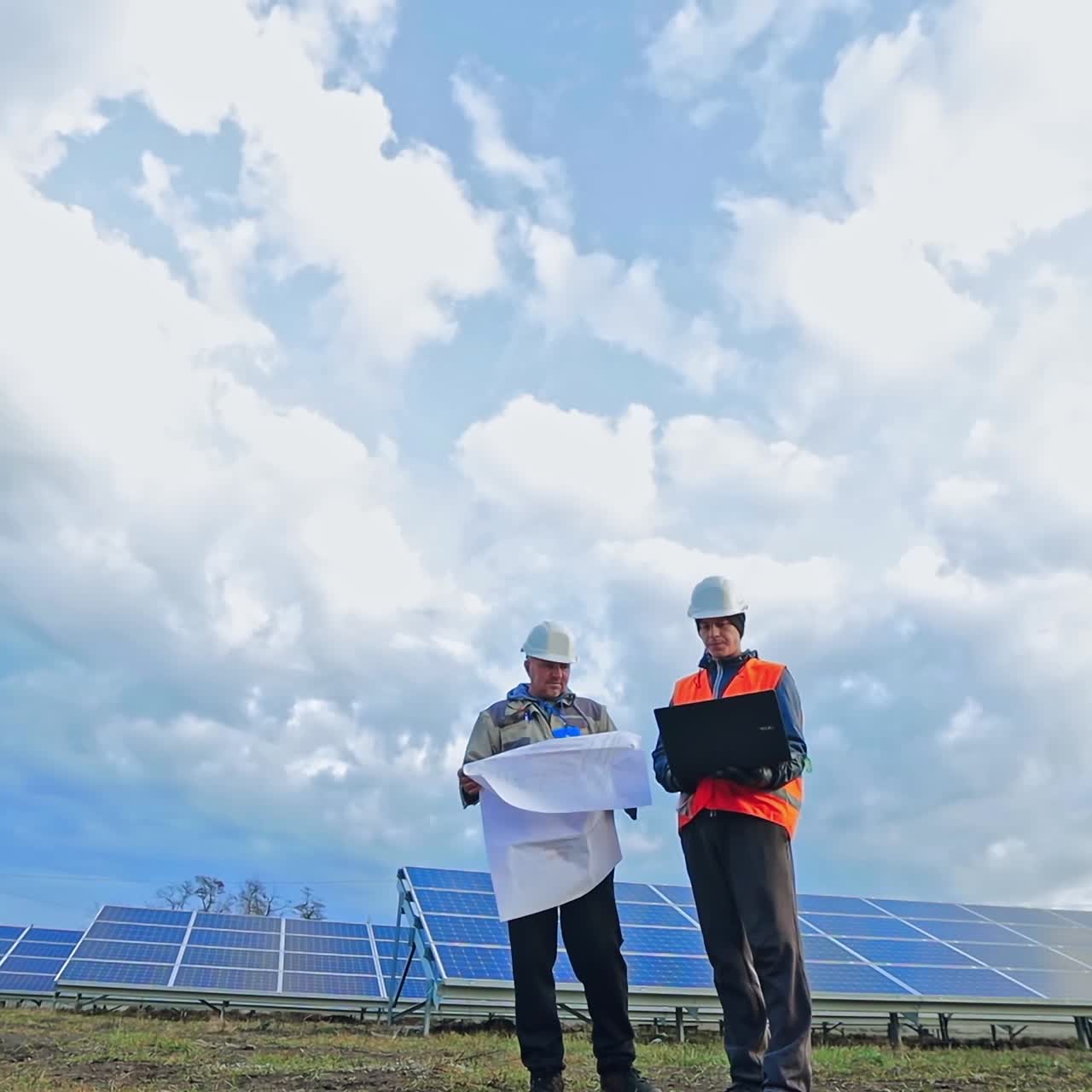 Engineers with laptop and project plan on the solar farm. Workers in special uniform standing among the solar panels and discuss the installation of sunny batteries.