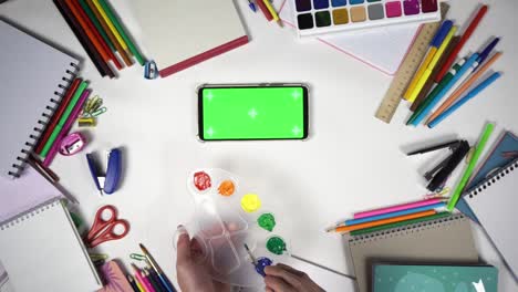 man is learning to draw using palette with colours and brush watching online lesson on a smartphone