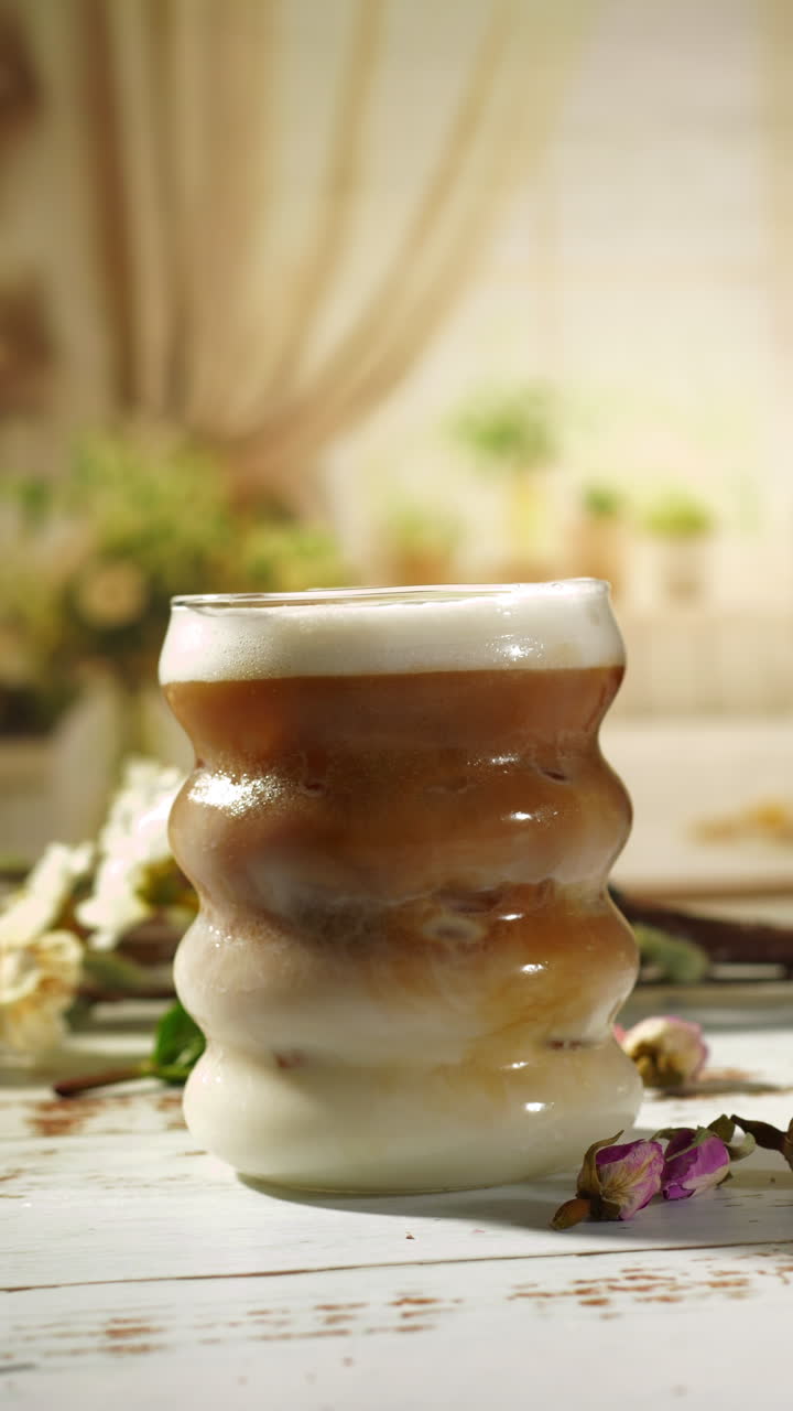 Camera tilts down as rich coffee is poured from a glass carafe into a bubble-shaped glass topped with creamy oat milk foam, set on a whitewashed wooden table strewn with dried blossoms.