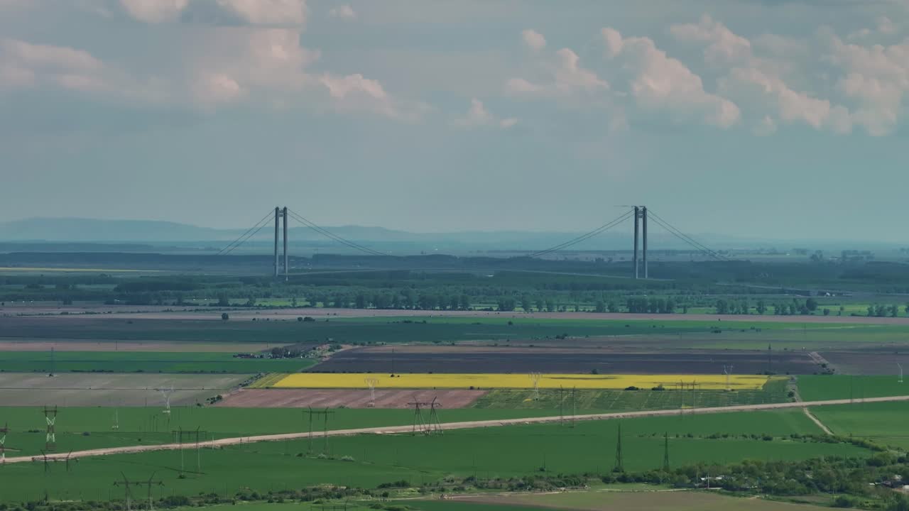 Luchtopname op afstand van een lange hangbrug, omkaderd door groene landbouwvelden en een bewolkte blauwe lucht