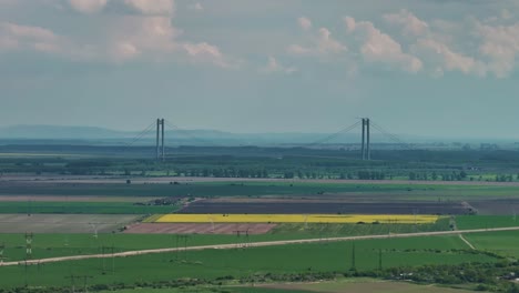 aerial distant shot of a long suspended bridge framed by green agricultural fields and a cloudy blue sky