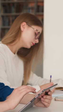 woman surfs internet on tablet in library hall. lady searches information on internet near girl friend writing homework at table. education for students