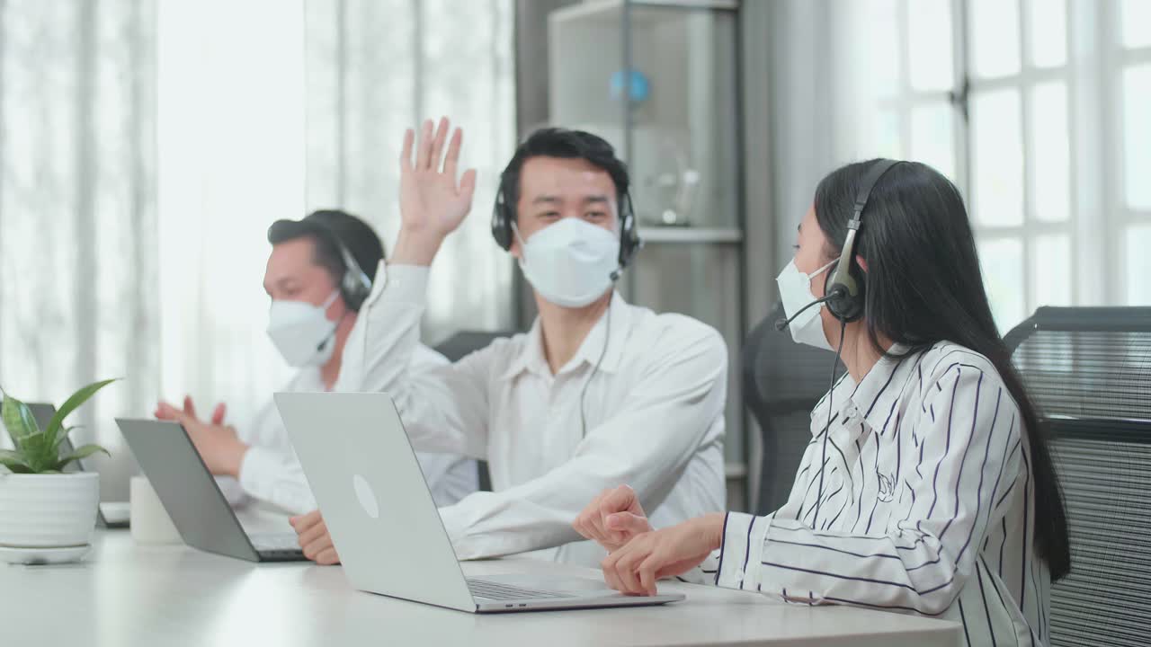 Three Asian Call Center Agents Wearing Headsets And Masks Sitting On The Chairs And Being Happy Due To Success Working With Computers At The Office