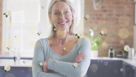 Animación-De-Puntos-De-Luz-Y-Flores-Sobre-Una-Feliz-Mujer-Caucásica-Sonriendo-En-La-Cocina