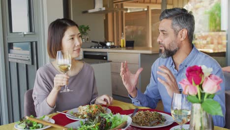 happy diverse couple sitting at table in dining room, eating dinner and drinking wine