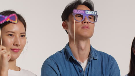 Side-View-Of-Group-Of-Young-Friends-In-Front-Of-White-Studio-Background-Having-Fun-Posing-For-Photo-Booth-Style-Portraits-With-Props