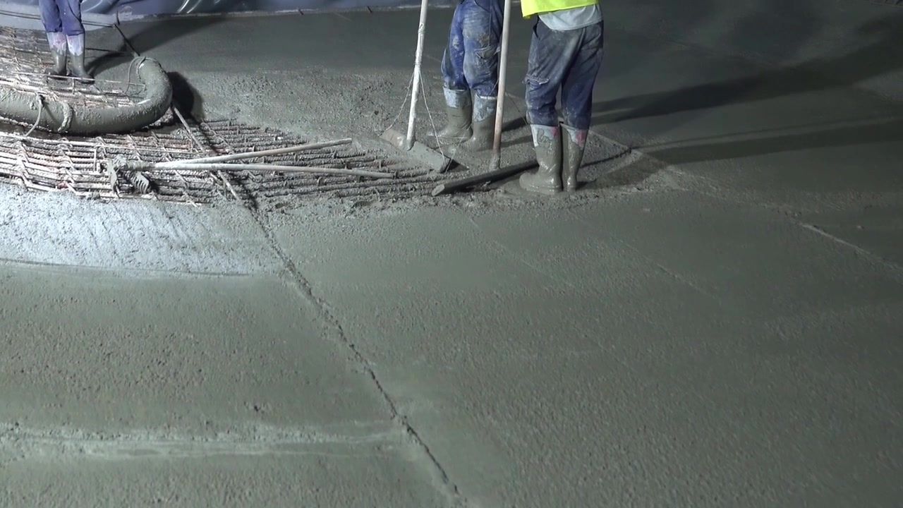 Bygningsarbeidere i vernestøvler jevner ut fersk betong over et armeringsjernnett av stål under et nattskift [kilde: AA_17.jpg]. Detaljbilde av industriell platehelling og anleggsdrift.