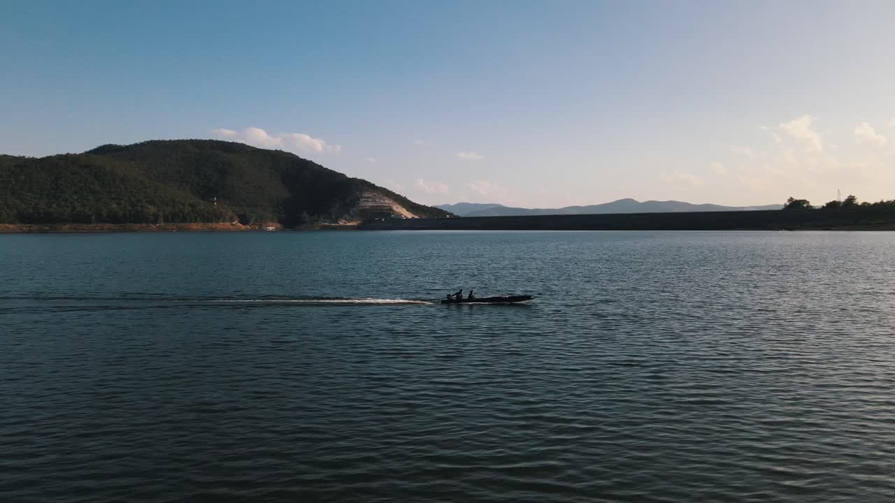 embarcación tailandesa tradicional larga y estrecha que viaja a gran velocidad sobre mae ngat somboon con la represa chon al fondo en un día soleado