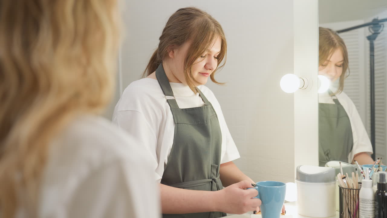 Beautician wearing gray apron holding cup of tea and saucer with gentle smile while approaching client inside beauty studio, creating warm and welcoming salon environment under soft indoor lighting