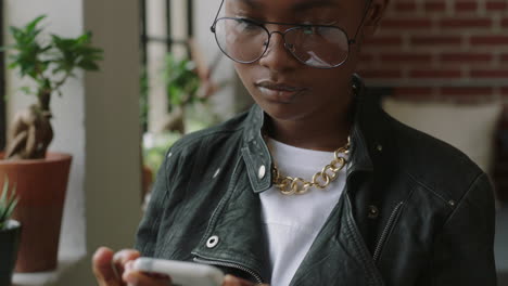 Elegante-Joven-Afroamericana-Usando-Un-Teléfono-Inteligente-En-Casa-Estudiante-Navegando-Mensajes-De-Redes-Sociales-Enviando-Mensajes-De-Texto-En-Un-Teléfono-Móvil-Disfrutando-Compartiendo-Un-Estilo-De-Vida-Moderno-En-Línea-Usando-Gafas