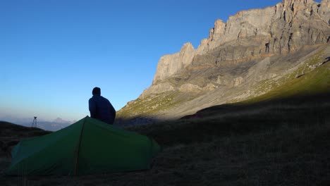 Hombre-Levantándose-Fuera-De-Su-Tienda-En-Una-Mañana-Fresca-Y-Mirando-Un-Acantilado-De-Roca-Iluminado-Por-El-Sol-En-Los-Alpes-Franceses