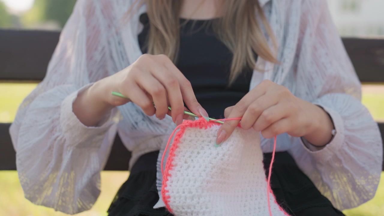 Primer plano de una mujer joven tejiendo a ganchillo con hilo rosa sobre tela al aire libre. La escena muestra manos trabajando con constancia, creando detalles de textura con luz natural suave contra un fondo exterior