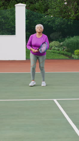 video vertical de una mujer biracial senior feliz jugando al tenis en una cancha de tenis