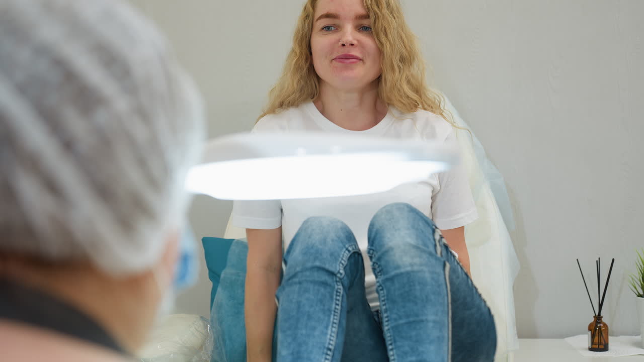 Client with blond hair sits on leather-covered chair, smiling and relaxed beside white salon lamp and air freshener in modern studio, ready for wellness or beauty treatment