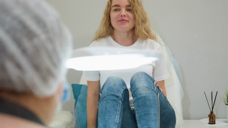 Client with blond hair sits on leather-covered chair, smiling and relaxed beside white salon lamp and air freshener in modern studio, ready for wellness or beauty treatment