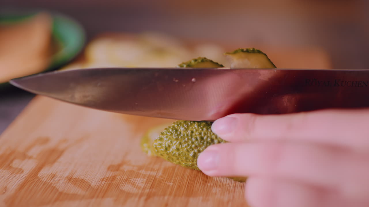 Close up of hand slicing fresh cucumber on wooden cutting board with knife, green vegetable texture visible, blurred cheese and bread in background creating kitchen preparation atmosphere
