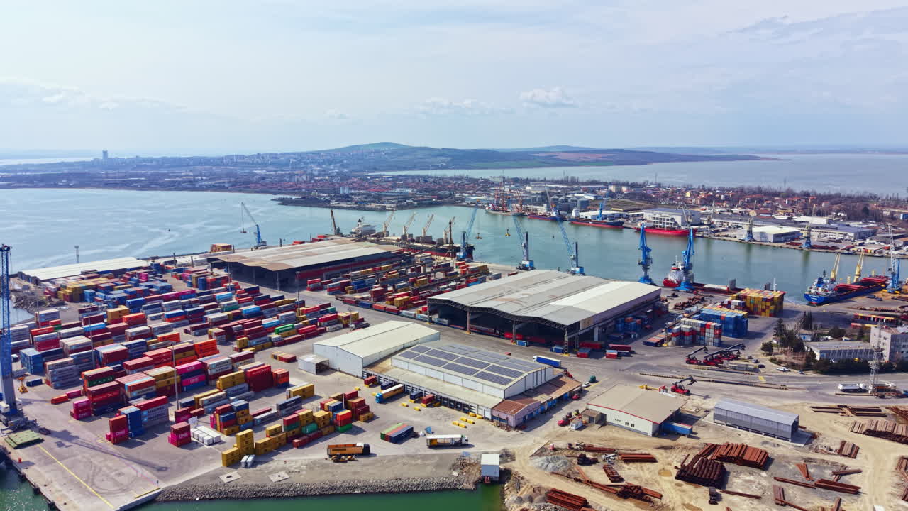 View of a busy shipping port with many containers and cranes near the water during daytime with ships and industrial area in the background