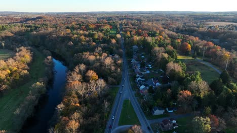 Aerial-view-of-a-road-by-a-river,-flanked-by-autumn-trees-and-houses-at-sunset