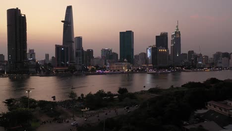 Ho-Chi-Minh-City,-Vietnam-iconic-Skyline-and-Saigon-river-waterfront-aerial-crane-shot-on-a-busy-evening-featuring-key-buildings-illuminated-against-colored-sky,-reflection-in-river-and-foreground