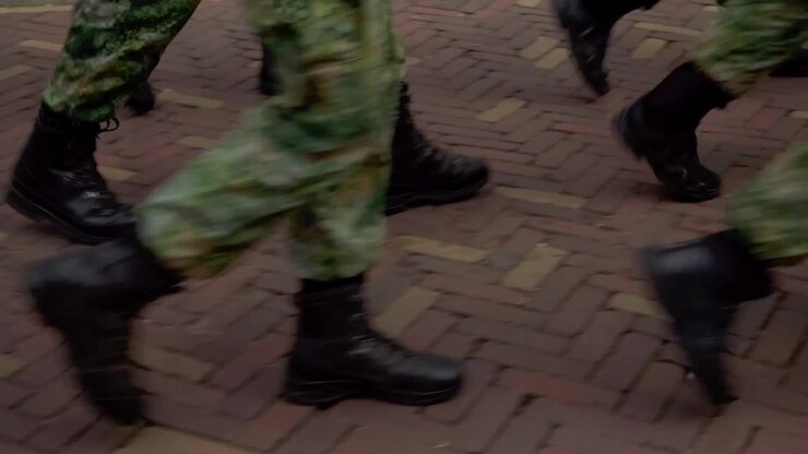 Primo piano dal basso di stivali militari e pantaloni mimetici che calpestano un tradizionale pavimento in mattoni mentre le forze armate marciano durante l'evento commemorativo Airborne a Oosterbeek