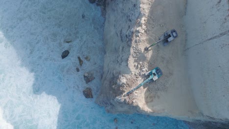 top down drone video of turquoise waves crashing against environmental destruction of the limestone cliffs near pura luhur uluwatu temple, bali indonesia with excavators dumping earth into ocean