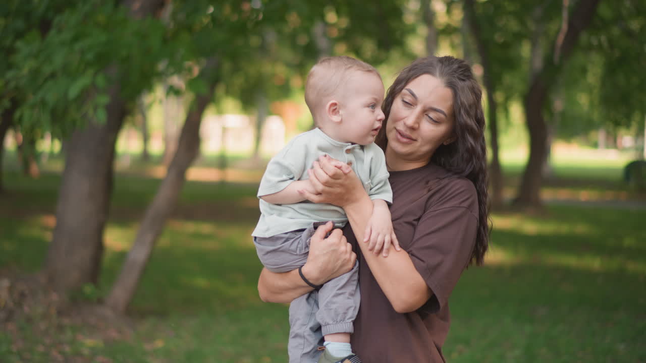 Madre affettuosa che tiene in braccio un bambino piccolo sotto gli alberi. La calda luce del sole avvolge madre e figlio che condividono affetto. Abbigliamento estivo casual mentre la madre abbraccia con amore il suo bambino sotto alberi ombrosi.