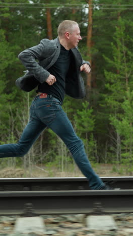A man wearing a grey suit jacket and blue jeans is captured mid-stride as he runs along railway tracks in a forested area, the lush greenery and electric poles in the background
