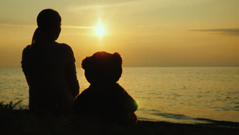 a lonely woman with a toy bear looks at the sunset over the sea sadness and loneliness concept