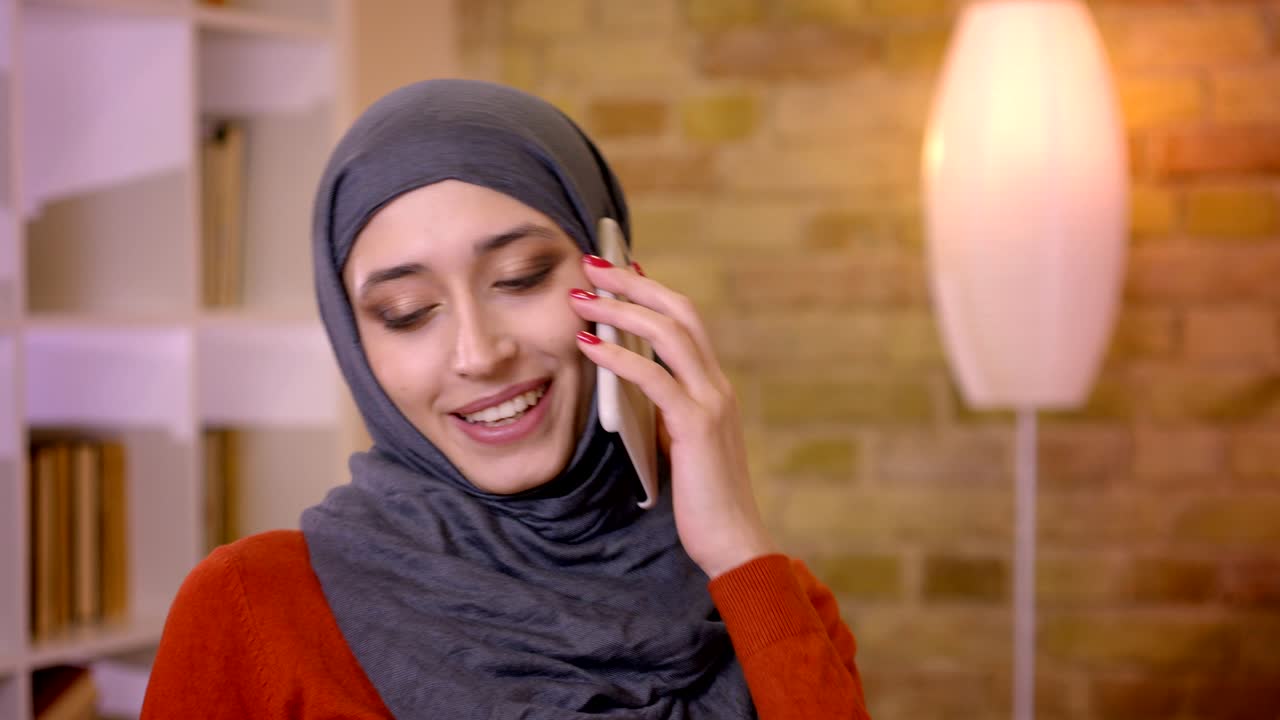 fotografía en primer plano de una joven empleada musulmana atractiva en hijab haciendo una llamada telefónica y sonriendo alegremente sentada frente a la computadora portátil en el interior de la oficina