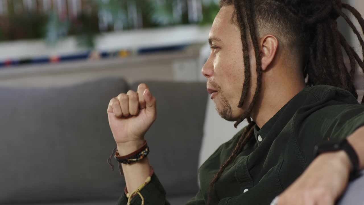 After camera pan, dreadlocked man is picking popcorn from bowl on lap and chewing while listening