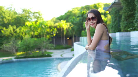 Pretty-Asian-woman-wearing-large-sunglasses-and-bathing-suit-relaxing-inside-a-pool-leaning-over-the-edge