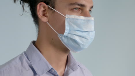 male with a man bun and smoldering eyes wearing a button down shirt puts on a covid-19 - coronavirus surgical mask on a grey background, side angle static shot