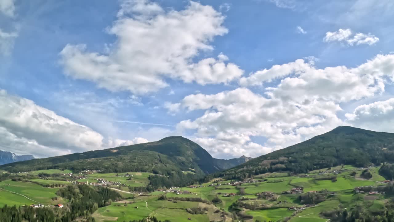 Vista panoramica di una lussureggiante valle alpina con fattorie e villaggi sparsi, pascoli verdi e dolci colline sotto un cielo punteggiato di nuvole, vicino alla strada per Innsbruck nell'Austria occidentale.