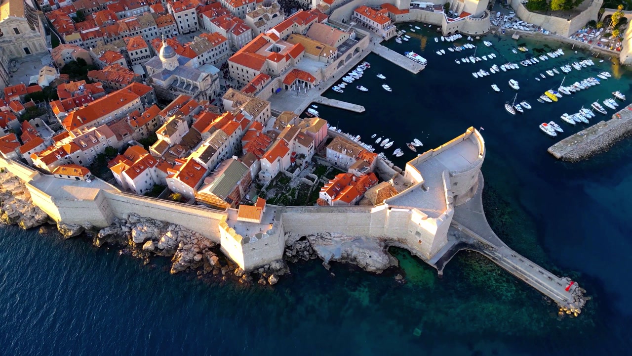 Stunning aerial drone footage of Dubrovnik's historic old town in Croatia, showcasing terracotta red-tiled roofs, ancient stone fortress walls extending into the deep blue Adriatic Sea.